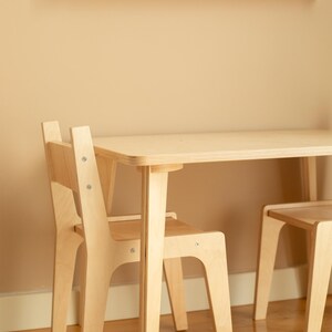 May include: A light-colored wooden children's table and chair set. The table has a rectangular top and four legs. Two chairs are visible, each with a backrest and seat. A wooden bookshelf with books is mounted on the wall above the table.