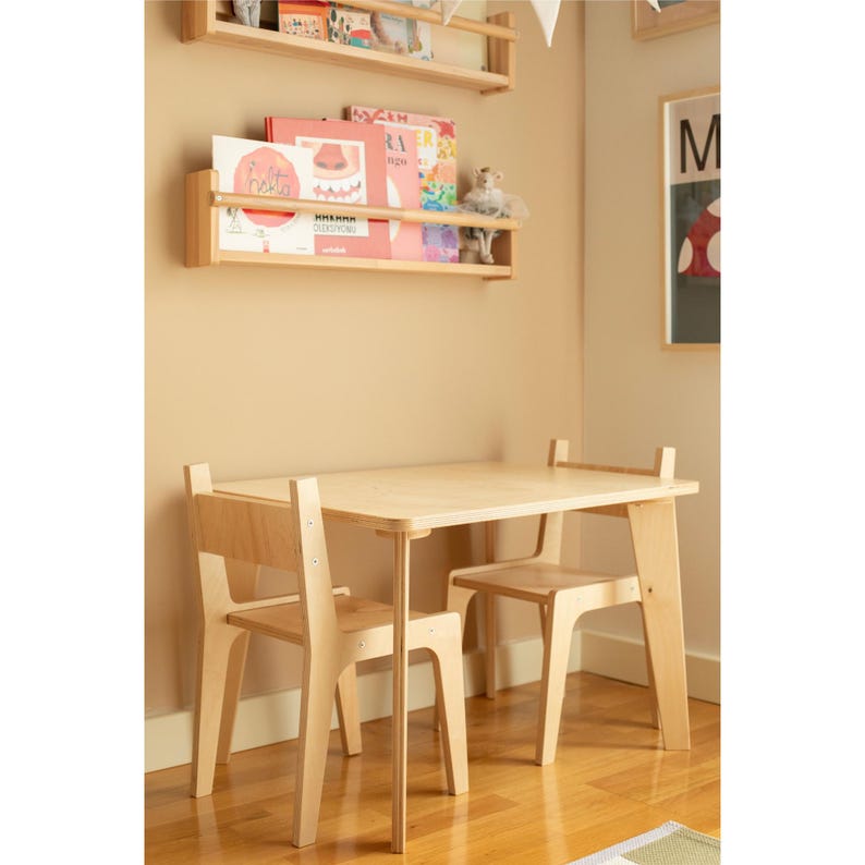 May include: A light-colored wooden children's table and two chairs set. The table has a rectangular top and straight legs. The chairs have a simple design with a backrest. The set is in a room with wooden shelves and books.