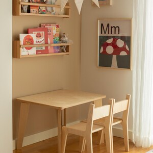 May include: A light wood children's table and two chairs set in a corner of a room. The table is rectangular with angled legs. A framed poster with a mushroom illustration hangs on the wall. Wooden shelves hold books and toys.