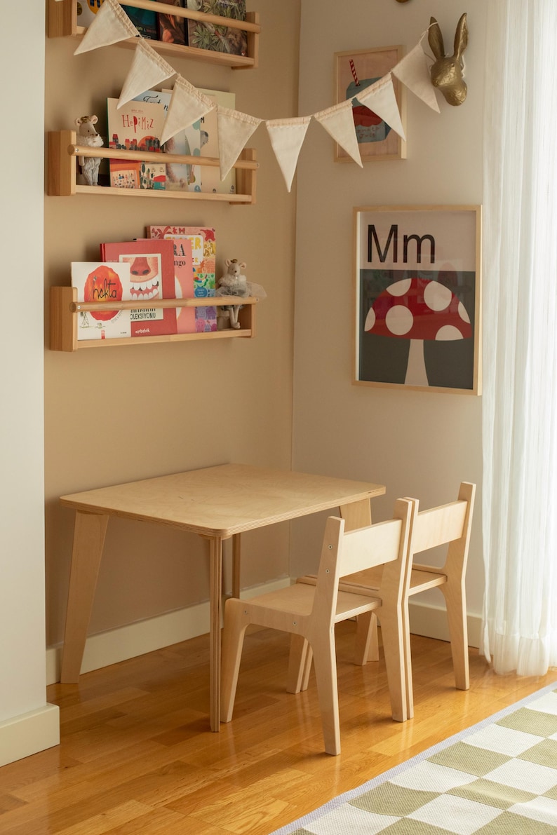 May include: A light wood children's table and two chairs set in a corner of a room. The table is rectangular with angled legs. A framed poster with a mushroom illustration hangs on the wall. Wooden shelves hold books and toys.