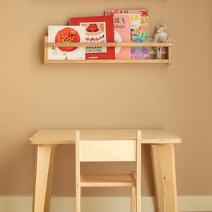 May include: A light-colored wooden children's desk and chair set. Two wooden shelves hold books and stuffed animals. A banner with triangular flags hangs above the shelves. The desk and chair are made of light-colored wood.