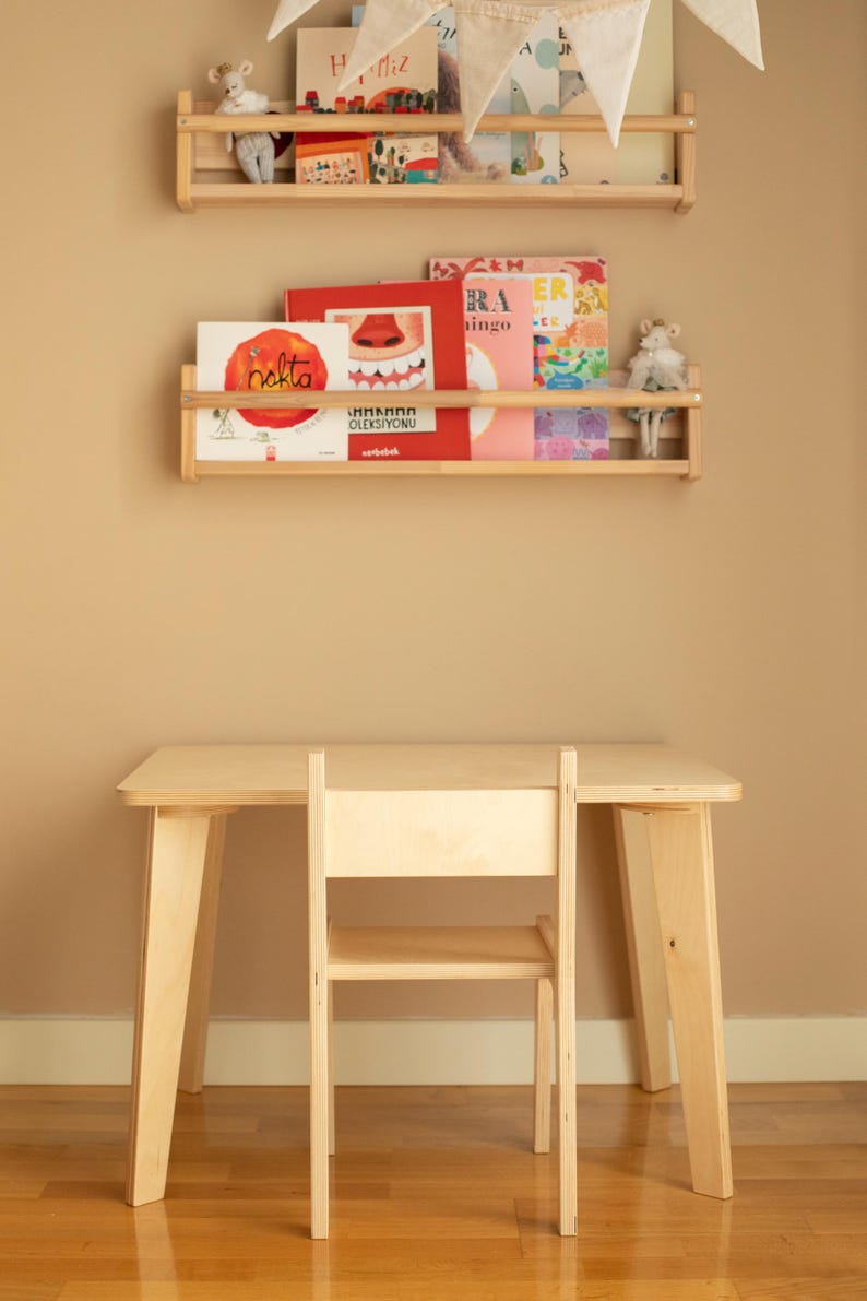 May include: A light-colored wooden children's desk and chair set. Two wooden shelves hold books and stuffed animals. A banner with triangular flags hangs above the shelves. The desk and chair are made of light-colored wood.
