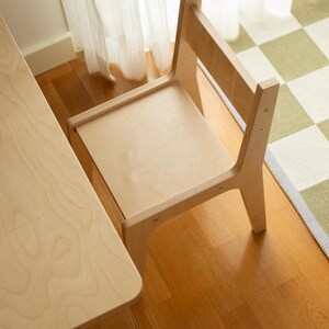 May include: A light-colored wooden chair and table set. The chair has a simple design with a backrest and a seat. The table is rectangular. The set is placed on a hardwood floor, with a checkered rug and sheer curtains in the background.