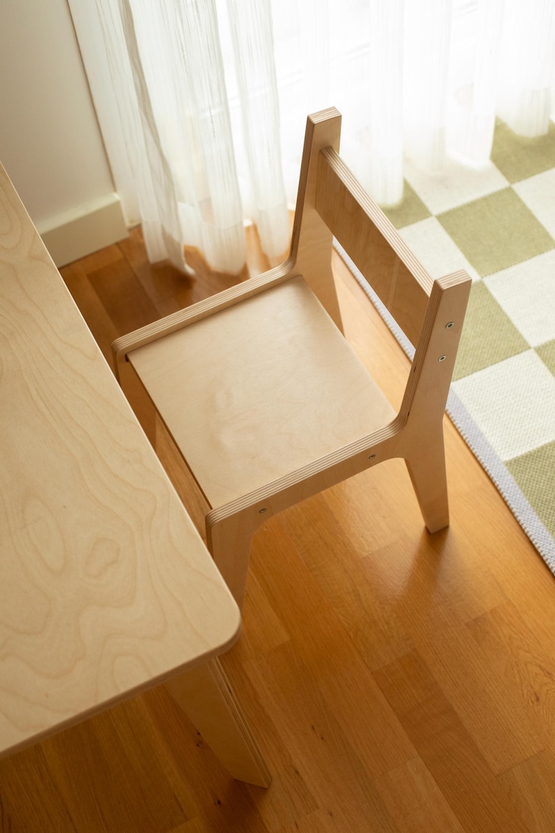 May include: A light-colored wooden chair and table set. The chair has a simple design with a backrest and a seat. The table is rectangular. The set is placed on a hardwood floor, with a checkered rug and sheer curtains in the background.