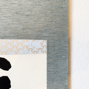 May include: Close-up of a framed artwork with a textured, gray-blue border. The artwork features a cream-colored paper with black brushstrokes and a decorative patterned border in gold and light blue.