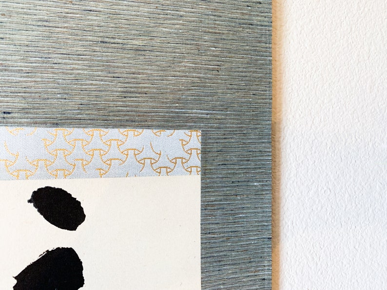 May include: Close-up of a framed artwork with a textured, gray-blue border. The artwork features a cream-colored paper with black brushstrokes and a decorative patterned border in gold and light blue.