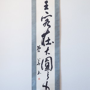 May include: A vertical Japanese scroll with calligraphy in black ink on white paper. The scroll is framed by a gray-green fabric. The calligraphy includes several characters and a red seal. A black rod is at the bottom.