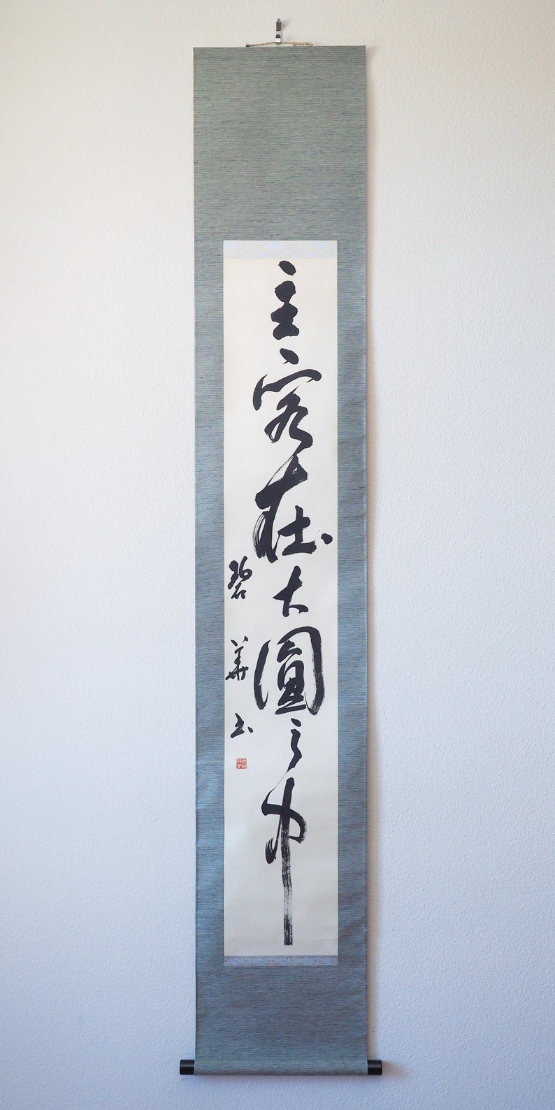 May include: A vertical Japanese scroll with calligraphy in black ink on white paper. The scroll is framed by a gray-green fabric. The calligraphy includes several characters and a red seal. A black rod is at the bottom.