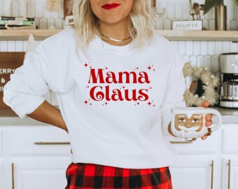 Op de afbeelding: Witte trui met rode tekst die "Mama Claus" zegt met rode glittersterren rond de tekst. De persoon draagt een rood-zwarte geruite rok.