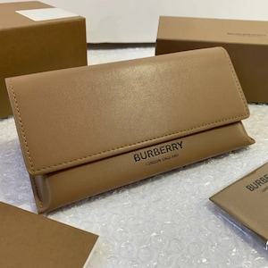 May include: A brown leather Burberry glasses case with the text "Burberry London England" on the front. The case is closed and sitting on a white surface.