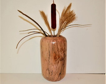 May include: A cylindrical wooden vase with a natural light brown grain and dark, irregular streaks. The vase contains a decorative arrangement of dried grasses and a single, prominent, dark brown, velvet-textured cattail plant stem.