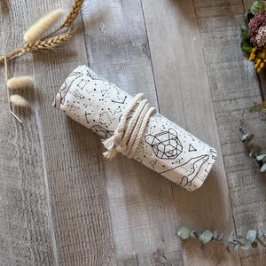 May include: Rolled white canvas wrap with a black constellation and geometric animal design, secured with a natural rope. The wrap is lying on a light wood surface, with dried plants and flowers in the background.