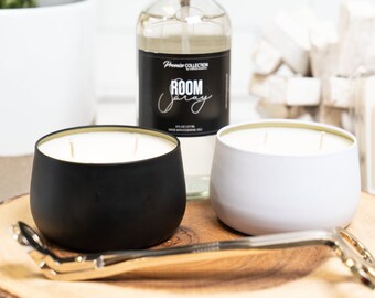 May include: Two candles in black and white metal containers with a gold rim. The candles are on a wooden surface with a clear glass bottle of room spray.