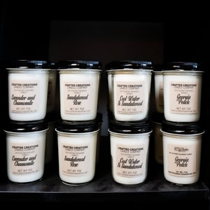 May include: A shelf display of nine soy coconut blended candles in glass jars with black lids. The candles are labeled with the brand name "Crafted Creations Candle Company" and various scents, including Lavender and Chamomile, Sandalwood Rose, Cool Water & Sandalwood, and Georgia Peach. Each candle is 7 ounces.