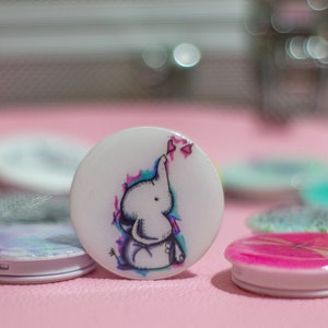 Puede incluir: Un agarre de teléfono blanco con un diseño de acuarela colorido de un elefante soplando corazones rosas.