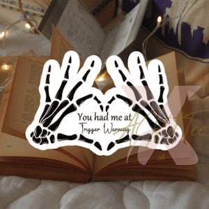 May include: A white sticker featuring two black skeleton hands forming a heart shape. The text "You had me at Trigger Warning" is in the center of the heart. The sticker is placed on an open book.