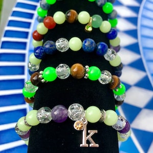 Puede incluir: Una pila de pulseras con cuentas de varios colores, incluyendo verde, azul, marrón y morado. Las pulseras están hechas con diferentes tipos de piedras, incluyendo ojo de tigre, lapislázuli y amatista. La pulsera inferior tiene un dije de plata con la letra 'K' en ella.