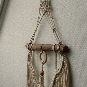 Puede incluir: Un tapiz de macram&eacute; de pared con una barra de madera y cuentas de madera. El tapiz tiene un borla en la parte inferior y est&aacute; hecho con cord&oacute;n de color natural.
