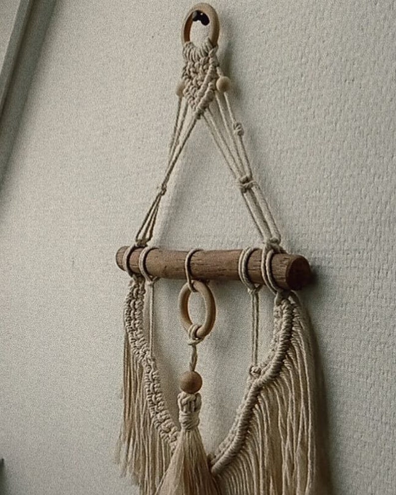 Puede incluir: Un tapiz de macram&eacute; de pared con una barra de madera y cuentas de madera. El tapiz tiene un borla en la parte inferior y est&aacute; hecho con cord&oacute;n de color natural.