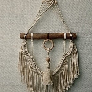 Puede incluir: Un tapiz de macram&eacute; de pared con una barra de madera y un borla. El tapiz est&aacute; hecho con cord&oacute;n de color natural y tiene un dise&ntilde;o geom&eacute;trico.