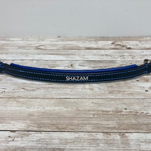 Peut inclure: Une sangle en cuir noir avec des coutures bleues et des ferrures argentées. La sangle porte le mot "SHAZAM" en relief.