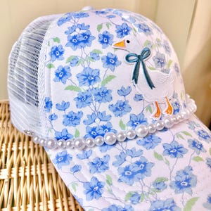 May include: White trucker hat with a floral pattern of blue flowers and a white mesh back. The hat features an embroidered goose with a teal bow and a pearl bead brim.