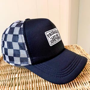 May include: Black trucker hat with a checkered pattern on the side and the text "PROBABLY - LATE FOR SOMETHING" on a rectangular patch. The hat is sitting on a woven basket.