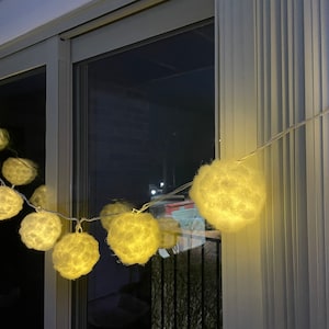 May include: A string of white fluffy ball lights with warm white LED bulbs. The lights are strung across a window and illuminate the room with a soft glow.