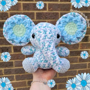 May include: A crocheted blue and white elephant with blue flower accents on the ears. The elephant is sitting on a hand.