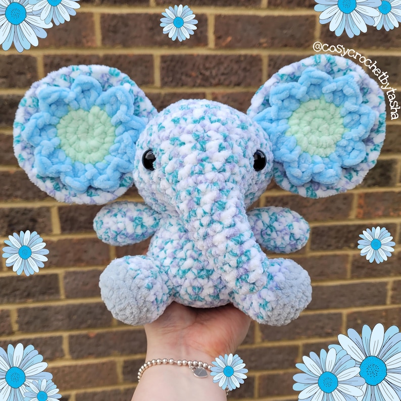 May include: A crocheted blue and white elephant with blue flower accents on the ears. The elephant is sitting on a hand.