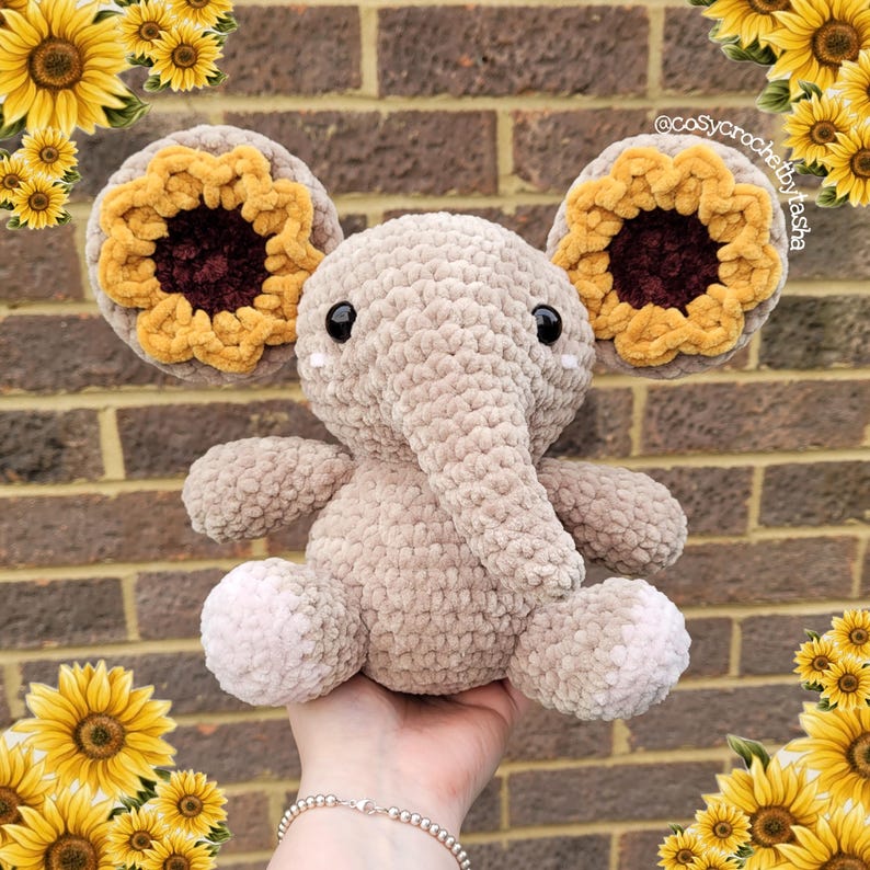 May include: A crocheted elephant plush toy with sunflower ears. The elephant is beige and has a long trunk. The sunflower ears are yellow with brown centers.