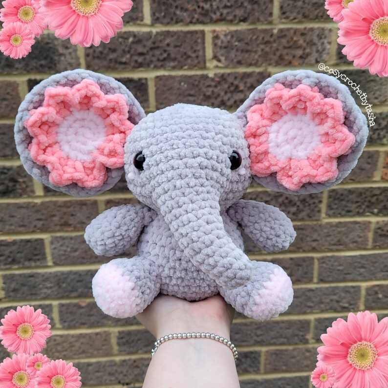 May include: A gray crocheted elephant plush toy with pink flower-shaped ears. The elephant has black eyes and a gray trunk. The toy is being held by a hand.