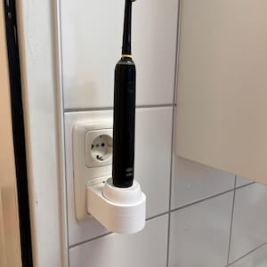 Könnte beinhalten: Eine schwarze elektrische Zahnbürste in einer weißen Ladestation, die an einer Steckdose angeschlossen ist. Der Bürstenkopf hat grüne und blaue Borsten. Die Einrichtung ist an einer weiß gefliesten Wand montiert.