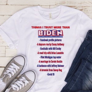 Puede incluir: Camiseta blanca con un gráfico azul y rojo que dice "Things I trust more than Biden" y enumera varios artículos. El gráfico está en una camiseta blanca con un par de jeans azules y zapatillas grises.