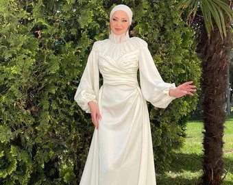 May include: A white satin wedding dress with long sleeves and a flowing train. The dress has a high neckline and a fitted bodice. The model is wearing a white headscarf.