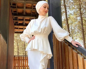 May include: A white satin dress with a ruffled skirt and a fitted bodice. The dress has long sleeves and a high neckline. The dress is worn by a person standing on a wooden staircase.