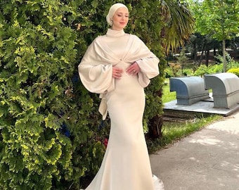 May include: A cream-colored formal gown with a high neckline and a flowing mermaid skirt. The dress features a cape-like top with wide sleeves. The person is wearing a matching head covering. The background includes green foliage and a paved walkway.