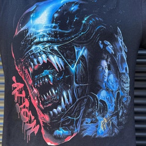 Könnte beinhalten: Schwarzes T-Shirt mit einem Grafikdruck eines außerirdischen Wesens mit scharfen Zähnen und leuchtend blauen Augen. Die Kreatur ist teilweise von einer menschlichen Figur mit einem ängstlichen Ausdruck verdeckt. Der Text "Alien" ist in Rot auf dem Shirt gedruckt.