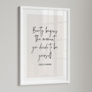 May include: A framed print with the quote "Beauty begins the moment you decide to be yourself" by Coco Chanel. The text is in a handwritten style, in black, against a light beige background, within a white frame.