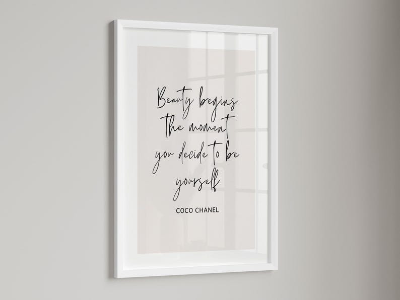 May include: A framed print with the quote "Beauty begins the moment you decide to be yourself" by Coco Chanel. The text is in a handwritten style, in black, against a light beige background, within a white frame.