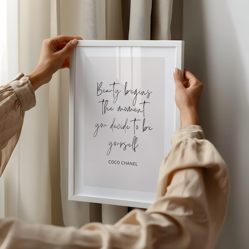 May include: A framed print with the quote "Beauty begins the moment you decide to be yourself" by Coco Chanel. The text is in a handwritten style, in black ink, on a white background. The frame is white.