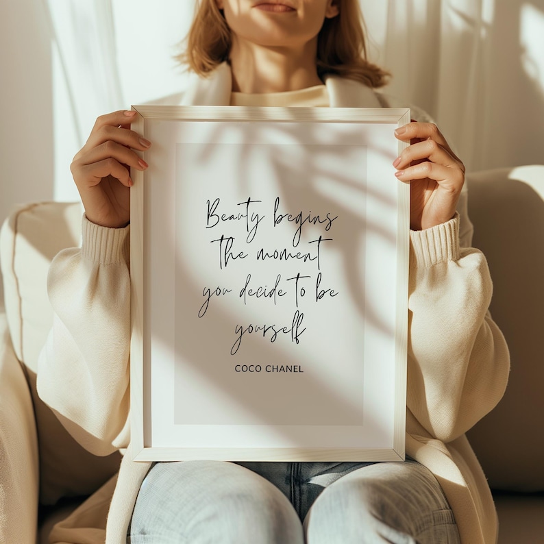 May include: A framed print with the quote "Beauty begins the moment you decide to be yourself" by Coco Chanel. The text is in a stylish script font on a white background, encased in a white frame.
