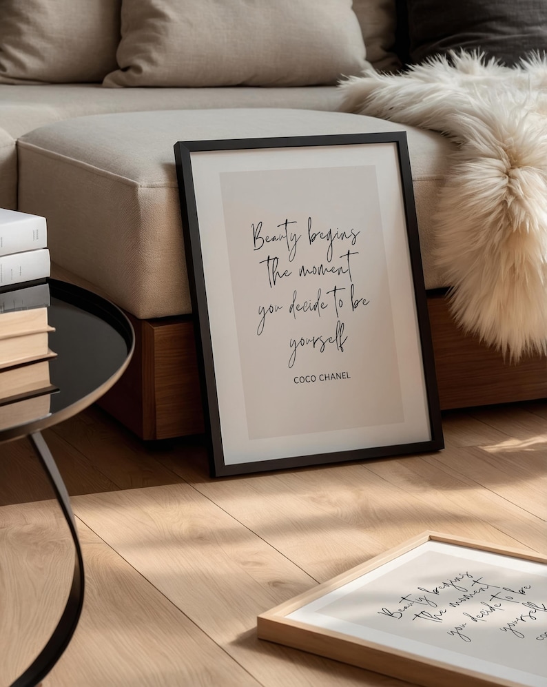 May include: A framed print with the quote "Beauty begins the moment you decide to be yourself" by Coco Chanel. The black framed print is leaning against a wall, with another framed print in the foreground. The background includes a sofa and a fluffy white throw.