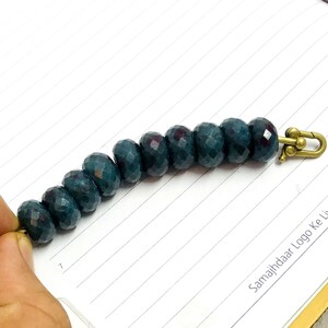 Könnte beinhalten: Ein Armband bestehend aus neun facettierten, dunkelblaugrünen runden Perlen, die in einer Reihe angeordnet sind. Die Perlen haben eine reflektierende, mehrfach facettierte Oberfläche. Ein messingfarbener Karabinerverschluss ist befestigt.