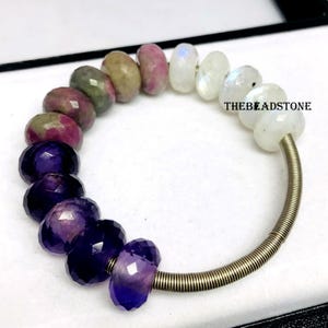 Könnte beinhalten: Ein Edelsteinarmband mit facettierten Perlen in Lila, Rosa, Grün und Weiß. Die Perlen sind auf einem gewickelten Metalldraht aufgefädelt. Der Text "THE BEADSTONE" ist auf der rechten Seite des Armbands sichtbar.