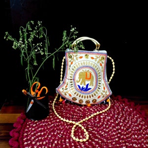 Könnte beinhalten: Eine weiße bestickte Handtasche mit einem goldenen Elefantenmotiv und einem Holzperlenriemen. Die Tasche liegt auf einem roten gehäkelten Teppich.