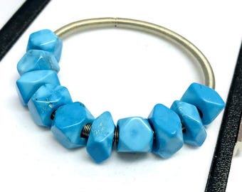 May include: A turquoise bracelet featuring irregular, faceted blue stones strung on a silver-colored, coiled metal band. The bracelet is displayed on a white surface, with a black border visible in the background. The stones have a polished finish.