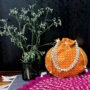 Puede incluir: Bolso de cordón naranja con adornos dorados y blancos. El bolso está decorado con lentejuelas y perlas. El bolso está sobre una alfombra de crochet rosa y blanca.