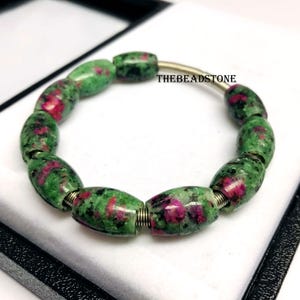 Könnte beinhalten: Ein Armband mit ovalen Perlen in Grün- und Rottönen. Die Perlen sind auf einem flexiblen Metalldraht aufgefädelt. Der Text "THEBEADSTONE" ist auf dem Draht zu sehen. Das Armband wird auf einer weißen Oberfläche präsentiert.