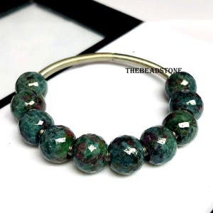 Pode incluir: Uma pulseira de contas redondas e facetadas, em tons de verde-azulado e bordô. As contas são enfiadas numa faixa de metal prateada e curva. O texto "THEBEADSTONE" é visível na faixa. As contas têm cerca de 1 cm de diâmetro.